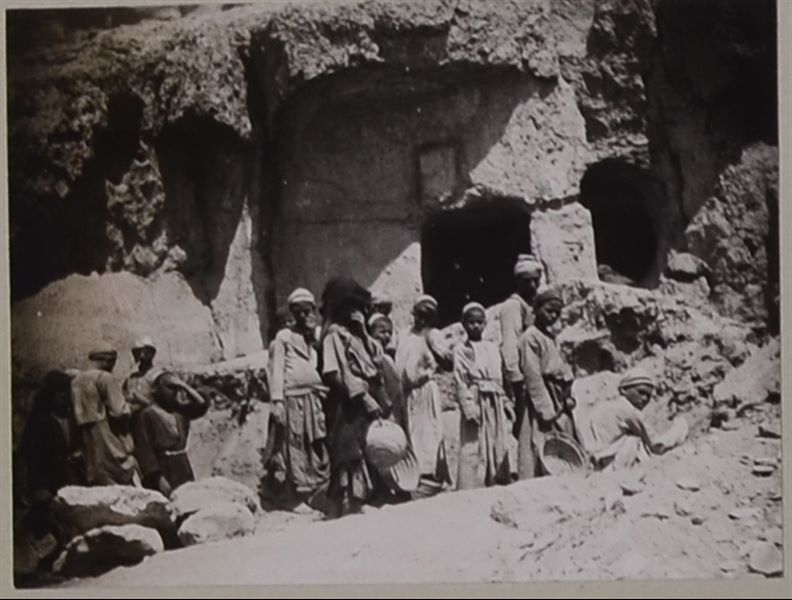 Shot taken during the cleaning and removal of debris from the external upper court of Governor Ibu's tomb, with several workers (adults and children) facing the camera lens. Behind them, the entrance to the part of the tomb excavated in the mountain can be seen. Photo album, Schiaparelli Excavations 