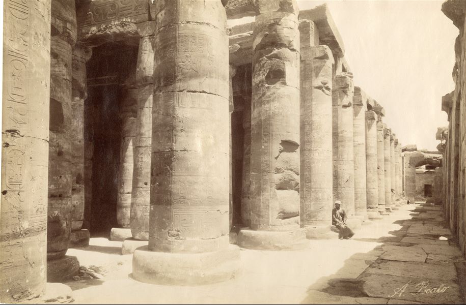 The picture shows part of the second hypostyle hall of the Temple of Pharaoh Seti I, the second ruler of the 19th dynasty, Abydos. In the photograph (taken from the north-west corner) the entrances to the chapels (right) are only just visible. At the bottom, the entrance to the "Hall of Nefertum and Ptah-Sokar". An egyptian is seated on the plinth of one of the columns. The author's signature is at the bottom right.  