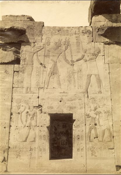 The photograph shows the reliefs from the back wall of the second hypostyle hall of the Temple of Seti I at Abydos. The ruler is portrayed together with some deities: Horus and Isis (above) and on either side of the entrance. The author's signature is visible at the bottom right.  