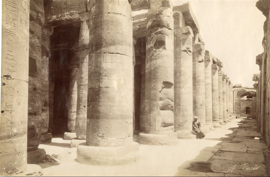 The picture shows part of the second hypostyle hall of the Temple of Pharaoh Seti I, the second ruler of the 19th dynasty, Abydos. In the photograph (taken from the north-west corner) the entrances to the chapels (right) are only just visible. At the bottom, the entrance to the "Hall of Nefertem and Ptah-Sokar". An egyptian is seated on the plinth of one of the columns. The author's signature is at the bottom right.  