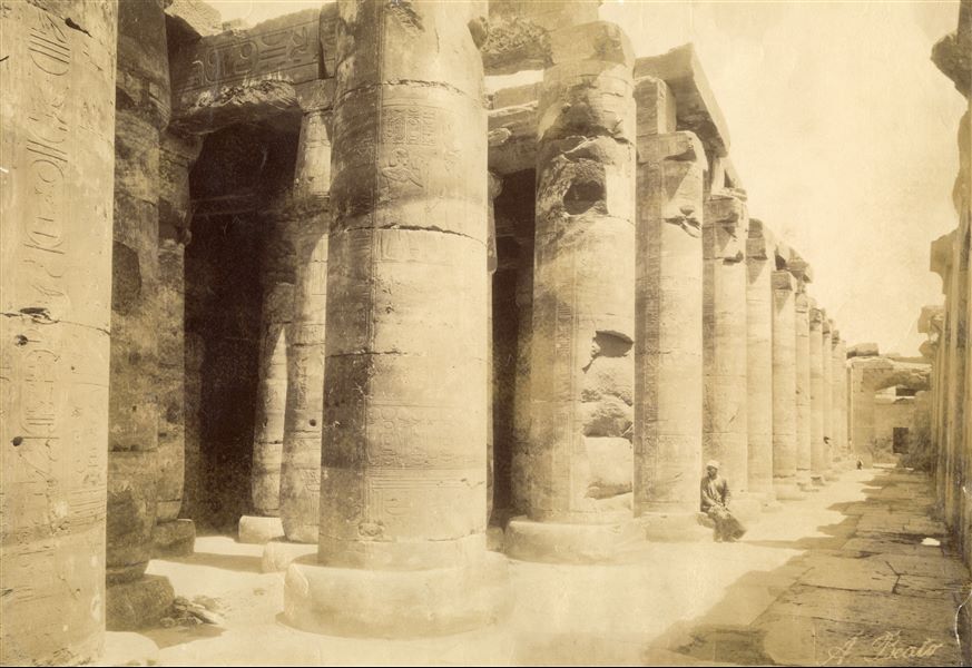 The photograph shows a cross-sectional view of the second hypostyle hall of the Temple of Seti I at Abydos, where the entrance to the chapels of the boats (right) and the "Hall of Nefertem and Ptah-Sokar" are visible. An egyptian sits at the base of the impressive columns, in the same position as image Inv02_002. The author's signature can be seen at lower right. 