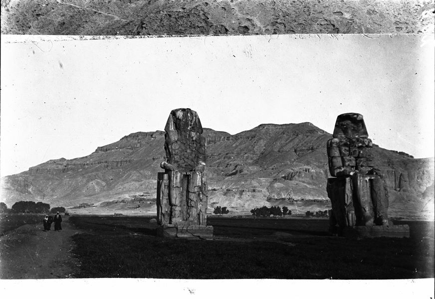 View of the so-called Colossi of Memnon. A pair of colossal statues depicting the pharaoh Amenhotep III seated on a throne. The Theban mountain is visible in the background. 